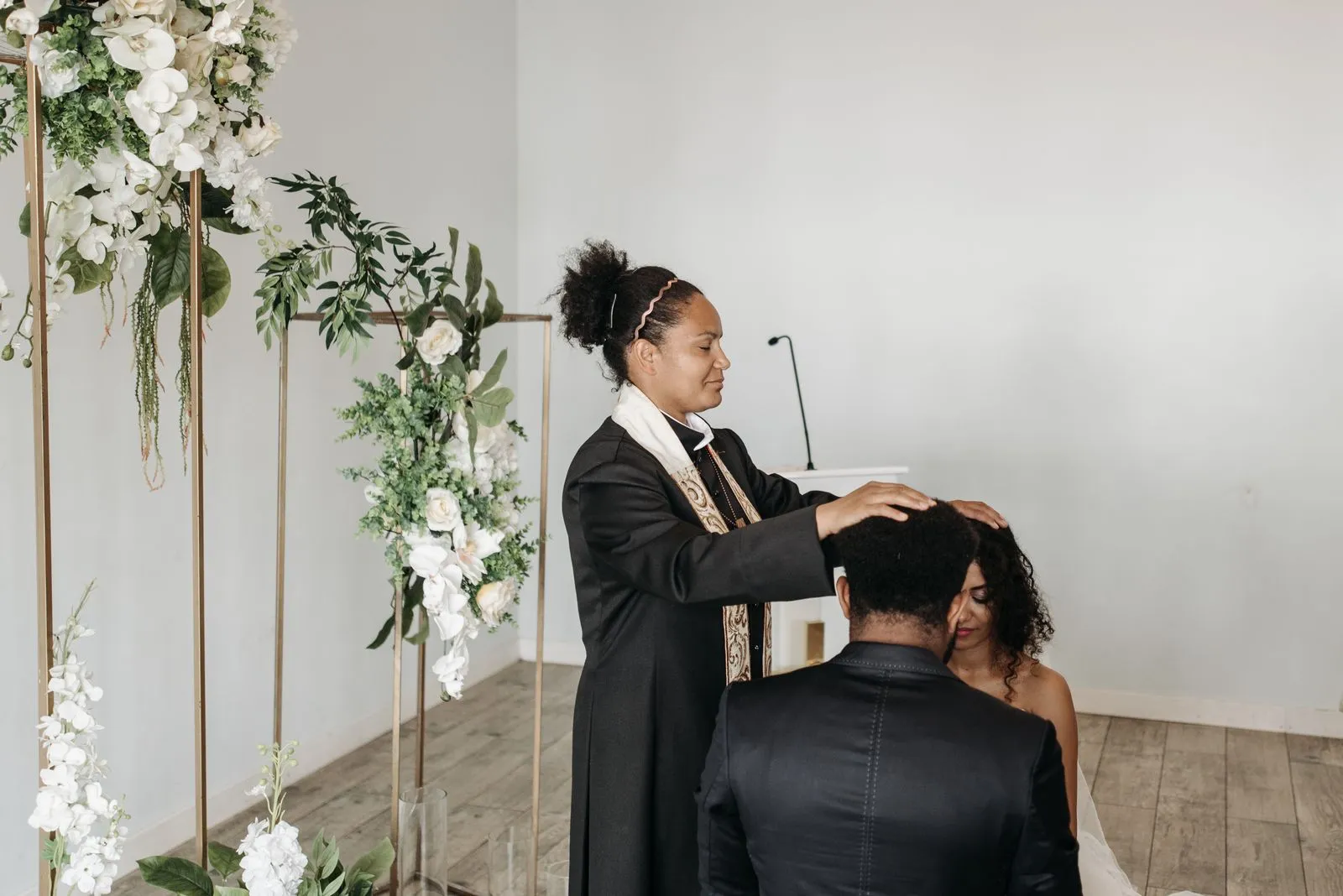 A wedding ceremony with a woman pastor blessing the bride and groom indoors.